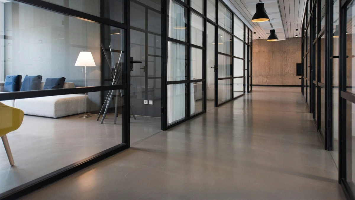 Das Bild zeigt einen modernen Büroflur mit Glaswänden und schwarzen Metallrahmen. Links befindet sich ein Besprechungsraum mit einem grauen Sofa, blauen Kissen und einer Stehlampe. Die Atmosphäre ist hell und minimalistisch, mit klaren Linien und schlichter Einrichtung.